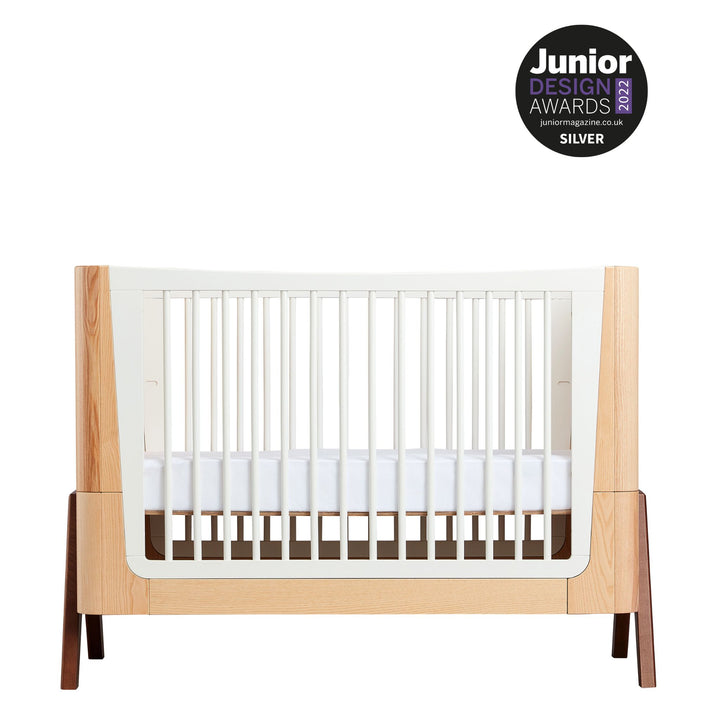 This image shows the stunning Hera Cot Bed in the Natural Ash and Walnut colour swatch on a white background with the Silver Junior Design Awards 2022 Logo displayed proudly in the top right-hand corner of the picture.